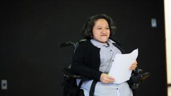 Photograph of young woman in a blue dress and a black sweater seated in a wheelchair holding a piece of paper on a stage.