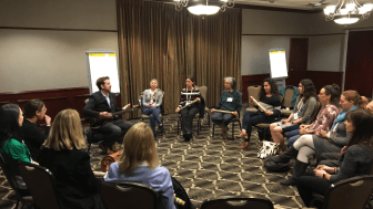 People sitting in a circle in a hotel conference room, Seneca Block holds a guitar and addresses the group