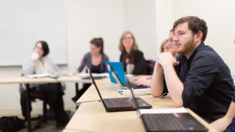Students in classroom off-campus, with focus on one boy student sitting with hands folded on the table with a laptop in front of him.