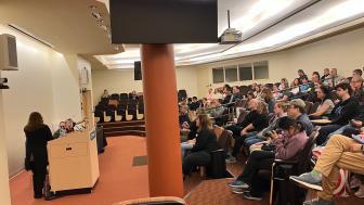 presentation in UHall amphitheater, Jen Thorell at podium, panelists at long table speaking to crowd seated in auditorium