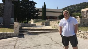 Tony Kecman in front of a museum in Croatia with stone buildings and green trees.