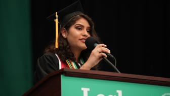 Woman on stage at Lesley Commencment behind the podium speaking into a microphone and wearing a black cap and gown.