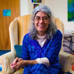 Photo of Jennie Kristel sitting in chair with hands folded in her lap, smiling at camera. She is wearing a royal blue shirt sitting on a neutral colored chair. 