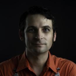 Headshot of Carl wearing an orange shirt with a black background