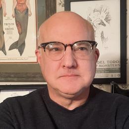 photo of faculty member John Rozum, wearing glasses, standing in front of wall art