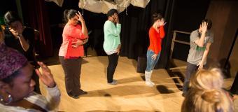 People in a circle on a stage during a drama therapy exercise stand with one hand up against their faces.