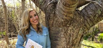 Katelyn Twardzik holding a children's book and leaning against a tree.
