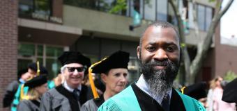 Matthew E. Henry wearing graduation robes with other people in the background. Standing outside.