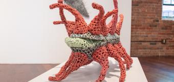 Artwork titled Germinate, made from red and green crocheted plastic. Appears to be standing on multiple legs with other tenticals come out of the top.