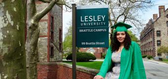 A photo of Natasha Gonzalez in a green cap in gown in front of the Lesley University sign on the South Campus. 