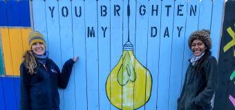 Two women stand in front of a panel of the mural/fence.
