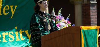President Steinmayer at the 2021 virtual commencement ceremony. She is standing in front of a podium on the quad on South Campus. There is a banner behind her that says "Lesley University."
