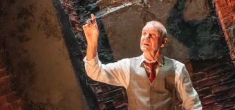 Man in a suit vest and pants holding up one hand, with a brick wall behidn him.