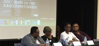 Four panelists are seated at a table with a black tablecloth and a screen with a PowerPoint can be seen behind them.