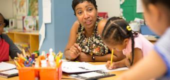 teacher sits in background assisting young students as they work on a project