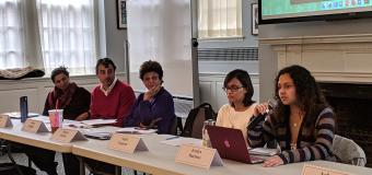 A panel of 5 speakers behind a table in Alumni Hall in front of a screen.