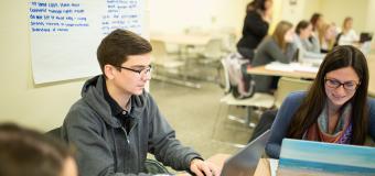 A group of graduate students at classroom tables with laptops