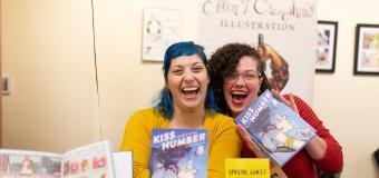 Ellen and Colleen sitting behind a table with their books on it, sitting next to each other laughing and holding their books up to their face
