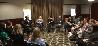 People sitting in a circle in a hotel conference room, Seneca Block holds a guitar and addresses the group