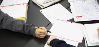 Close up of three pairs of hands at at a table, collaborating on a project with open notebooks filled with graphs and equations.