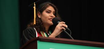 Woman on stage at Lesley Commencment behind the podium speaking into a microphone and wearing a black cap and gown.