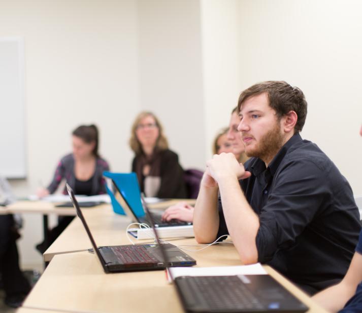 Students in classroom off-campus, with focus on one boy student sitting with hands folded on the table with a laptop in front of him.