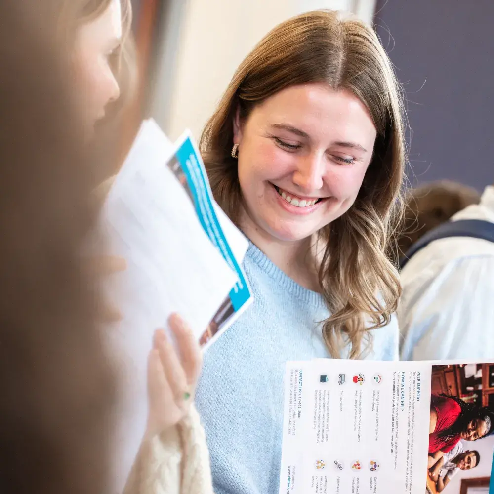 A smiling young woman with light brown hair, wearing a light blue sweater, looks down at a brochure she is holding. Her fingers are pointing to information on the page. Another person, seen from behind and out of focus, is partially visible on the left, also holding papers. The background is blurred, showing what appears to be an indoor setting with other people.