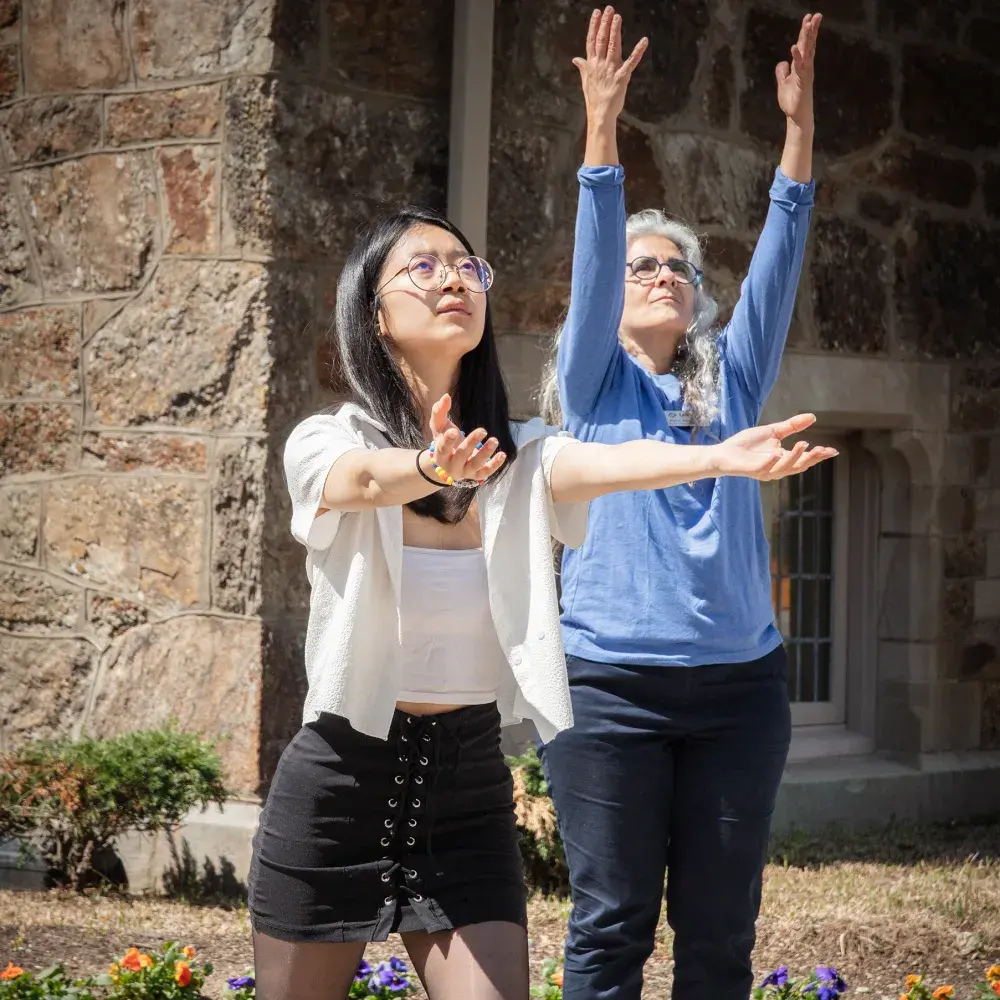 Two women demonstrate focus and mindfulness during an outdoor wellness class. The younger woman holds a dynamic forward-moving pose, while the older woman stands tall, reaching her hands toward the sky. Their different movements suggest a guided practice in a shared, sunny space.