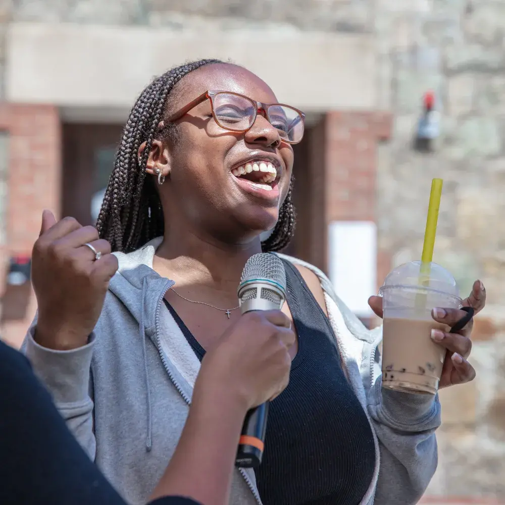 A close-up captures the vibrant energy of a young woman speaking at a wellness fair. Her face is full of joy as she talks into a microphone, holding a drink in her hand, suggesting she is sharing a positive experience or story.