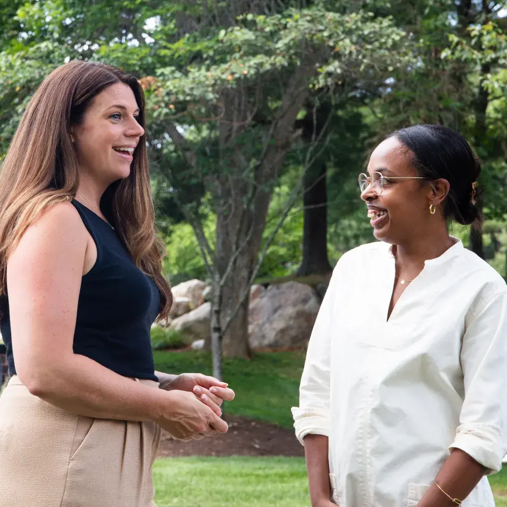 Two women are standing outdoors on a grassy area, smiling and looking at each other as if in conversation. The woman on the left has long brown hair, is wearing a black sleeveless top and beige pants. The woman on the right has dark skin, is wearing glasses and a white collared top. In the background, a blue sign with "125 HARTWELL AVENUE" and "Eliot" in white text is visible. Trees and some colorful flowers are also in the background.