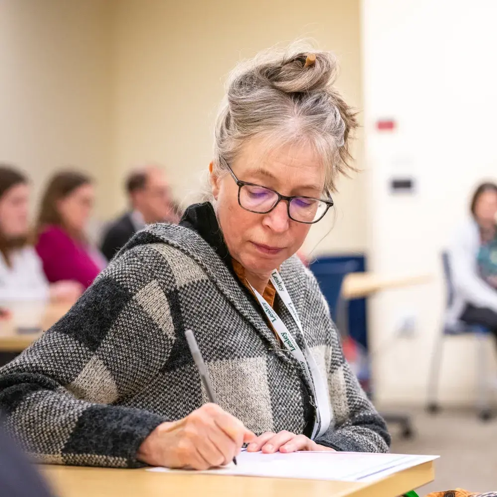 A woman with gray hair tied up in a messy bun wears glasses and a plaid wool sweater while writing on a piece of paper during a classroom workshop. She is seated at a desk and appears focused on the activity. A green Lesley lanyard hangs around her neck. Other participants can be seen blurred in the background, also engaged in writing. The setting is a well-lit educational environment, likely a seminar or conference.