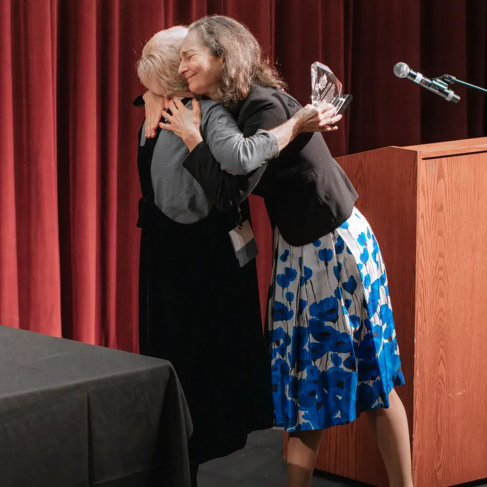 A woman with long, dark hair, wearing a black blazer and a white skirt with blue floral patterns, hugs another woman who has short, light-colored hair and is wearing a dark top and long dark skirt. The woman with dark hair is holding a clear, glass-like award in her left arm. To the right, there is a wooden podium with a microphone, and a black draped table with a microphone is on the left. A red curtain serves as the backdrop.
