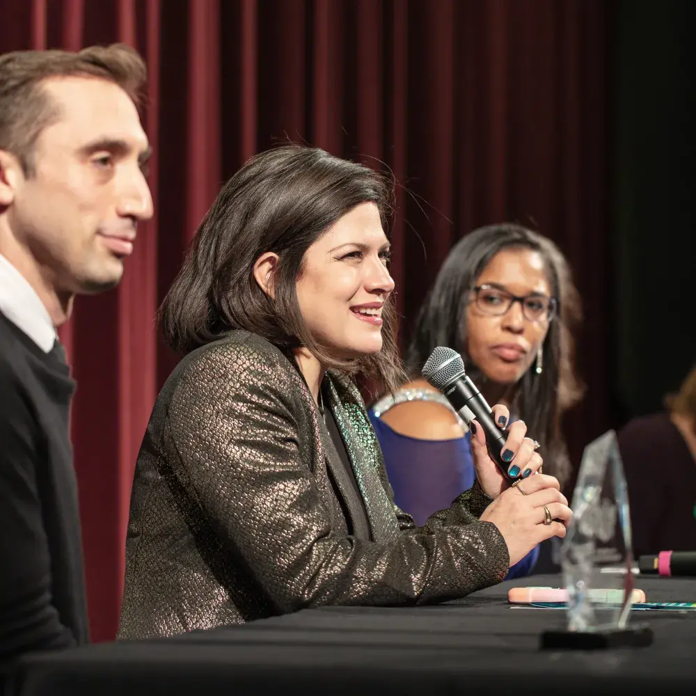 A smiling woman with dark hair and a shimmering gold jacket speaks into a microphone, sitting at a table with two other people partially visible. To her left, a man in a black sweater looks towards her. To her right, another woman with glasses and dark hair looks on. A dark red curtain is in the background, and a clear award or trophy is visible on the table to the right.