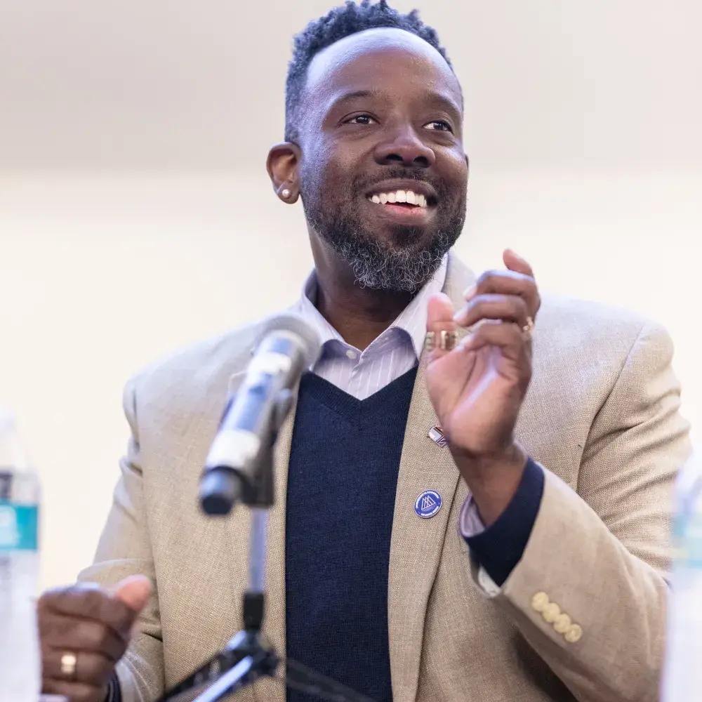 A professionally dressed man smiles warmly while speaking at a panel discussion. He wears a beige blazer over a navy sweater and collared shirt, with two pins on his lapel. He gestures with his hands as he speaks into a microphone on a stand. A water bottle sits on the table in front of him. The setting appears to be an indoor conference or seminar, likely an alumni or academic event.