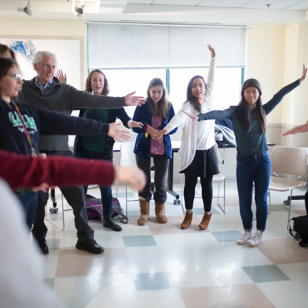 Participants in a workshop or class stand in a circle and engage in a group stretching exercise. Their synchronized movements and engaged expressions suggest a collaborative and energetic activity. The room is a typical classroom with a tiled floor and chairs moved to the side.