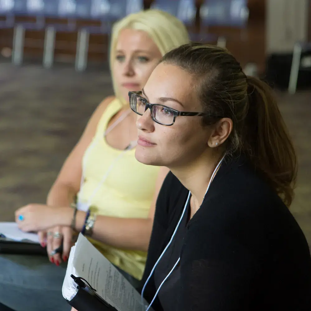 A close-up of a female attendee at a workshop, wearing glasses and a black shirt. Her rapt attention on an off-camera speaker conveys a sense of deep engagement and learning.