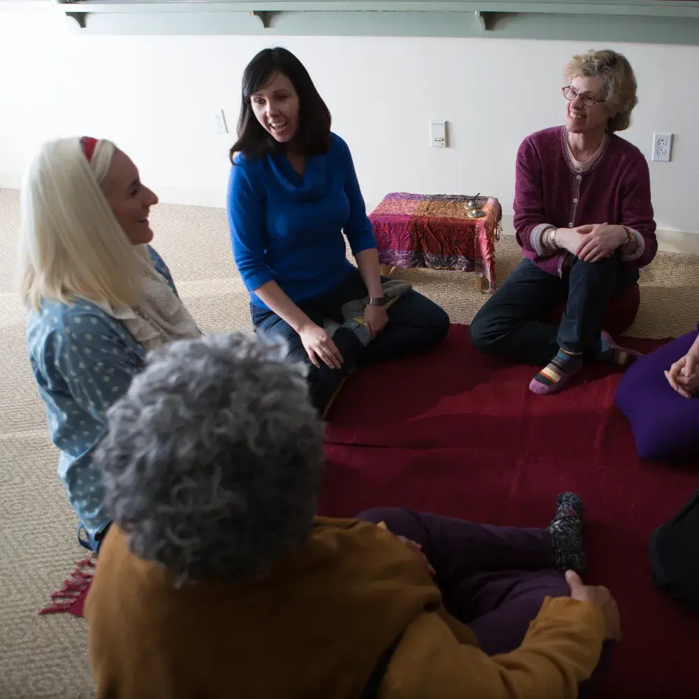 A group of six women sits in a circle, with six participants sitting on a red mat on the floor. Their engaged and comfortable postures create a sense of community and supportive conversation in a simple, well-lit room.