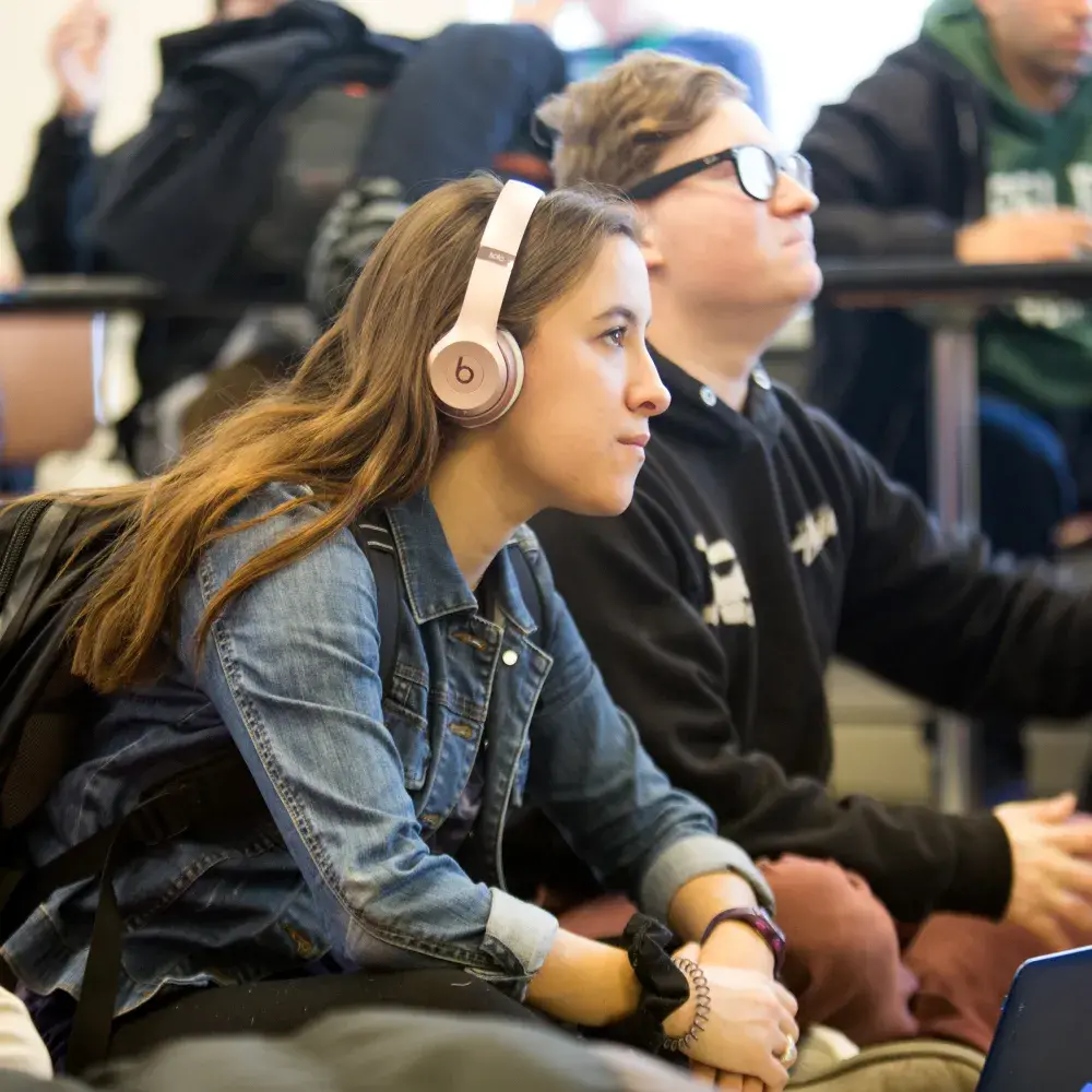 A young woman wearing pink Beats headphones and a denim jacket sits attentively next to a classmate in a busy classroom. She has a laptop open in front of her and a backpack on her shoulders. Around them, other students are engaged in the session, suggesting a collaborative and inclusive learning environment.