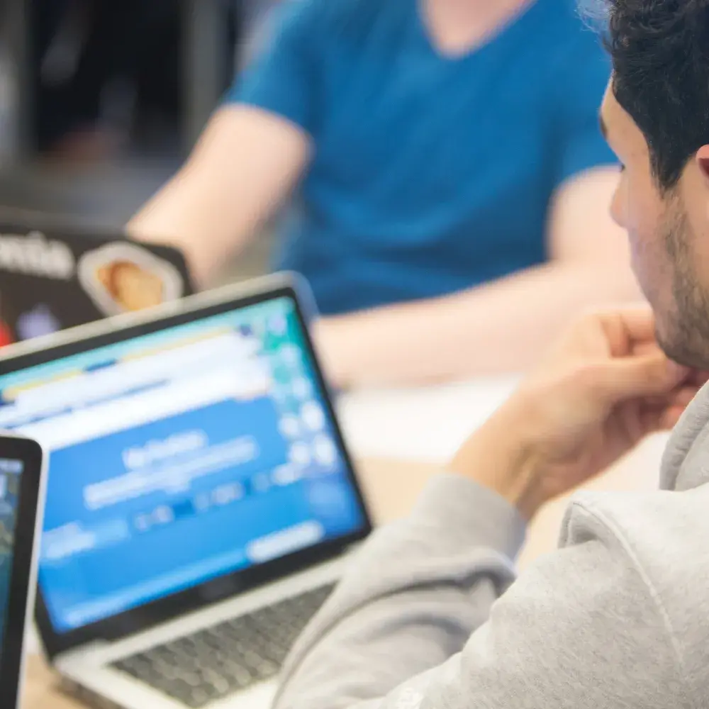 A student in a gray hoodie is seen from behind, sitting at a table and looking at a laptop screen. Several other laptops are on the table, one showing a desktop screen and another displaying a website. Another student in a blue shirt is seated across from them, slightly out of focus. The setting appears to be a classroom or study space.