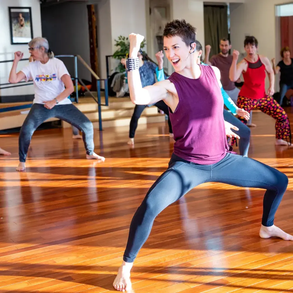 The vibrant energy of a group fitness class fills a sun-drenched studio. A charismatic instructor in the foreground strikes a powerful pose, leading a diverse group of people through an empowering, dance-based workout.