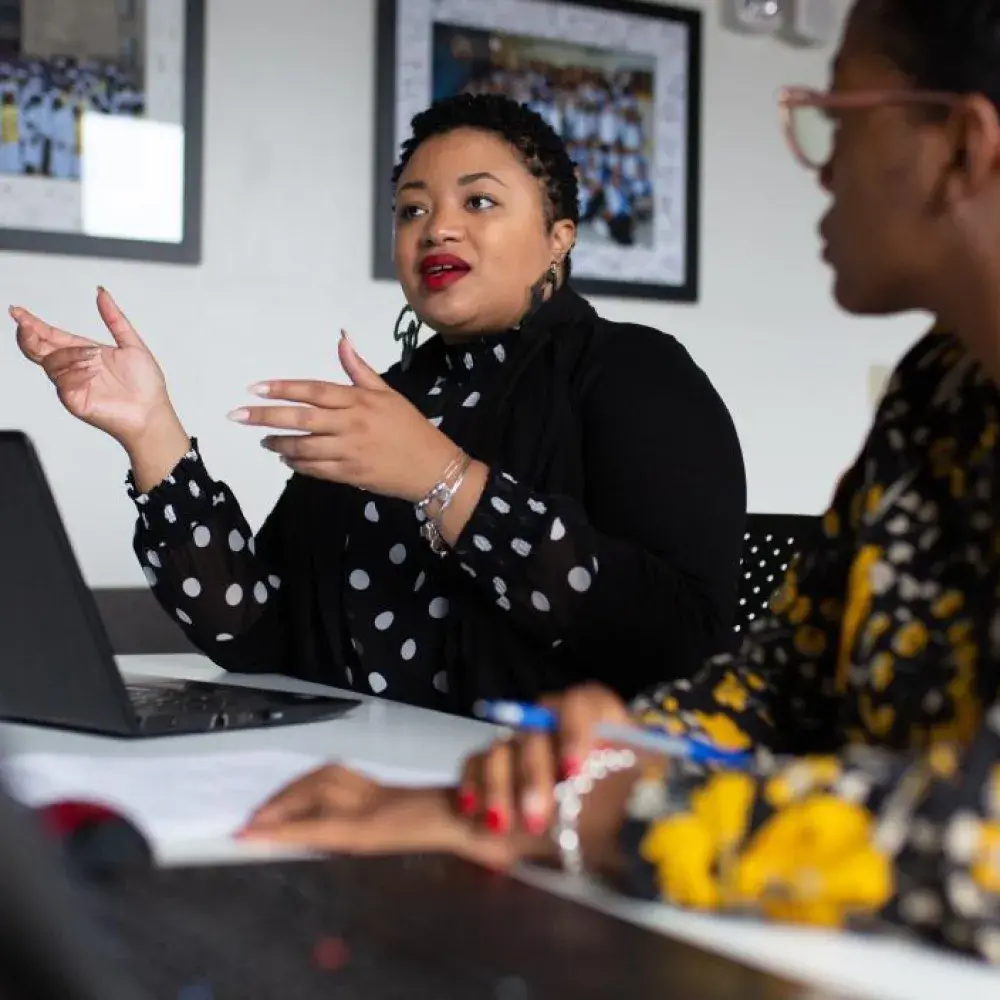  Two women are engaged in a professional discussion at a conference table with open laptops and documents. The woman in the center, wearing a black polka-dotted blouse and gesturing with her hands, is speaking confidently. The woman on the right, wearing a floral-patterned outfit, listens attentively. The setting appears to be an office or academic environment, with framed photos on the wall in the background.