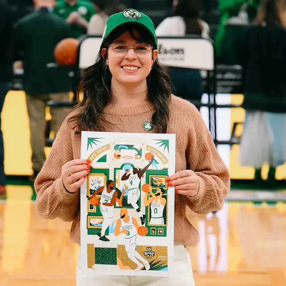 A smiling woman stands on a basketball court holding up a colorful illustrated poster featuring Boston Celtics players. She wears a green Celtics cap, glasses, a tan sweater, and a Celtics pin. People and activity are visible in the background, suggesting a game or event is taking place.