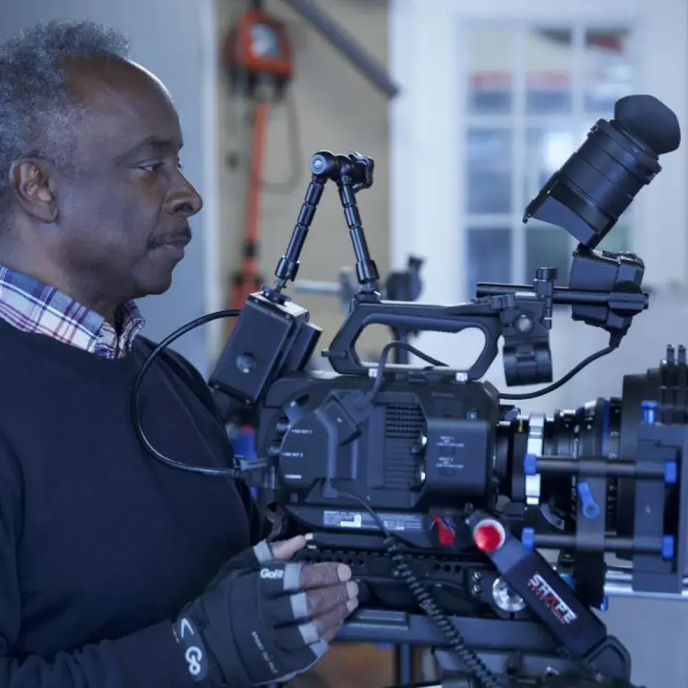 A man operates a professional video camera setup in a studio or workshop setting. He wears a dark sweater over a plaid shirt and fingerless gloves, focusing intently on the camera’s controls. The camera is equipped with various attachments, including a matte box, external monitor, and viewfinder. The background shows tools and equipment, suggesting a behind-the-scenes filming environment.