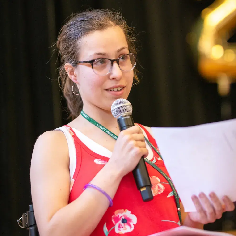 A young woman with glasses and a floral red dress speaks into a microphone while holding papers in her other hand. She wears a green lanyard with "Lesley University" printed on it and sits against a dark backdrop, with a gold balloon shaped like the number "2" slightly blurred in the background.