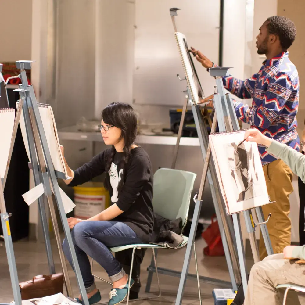 In a busy art studio, a diverse group of students of various ages participates in a life drawing class. They are scattered around the room, drawing on easels. In the foreground, a student holds up their charcoal portrait for critique, while an instructor looks on in the background.