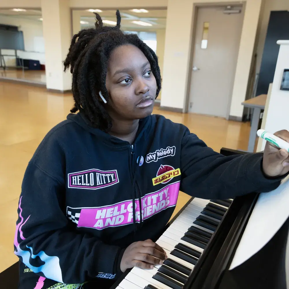 A young music student is captured in a moment of creative thought. They are sitting at a piano in a studio, holding a marker as if to notate music, with a pensive expression on their face.