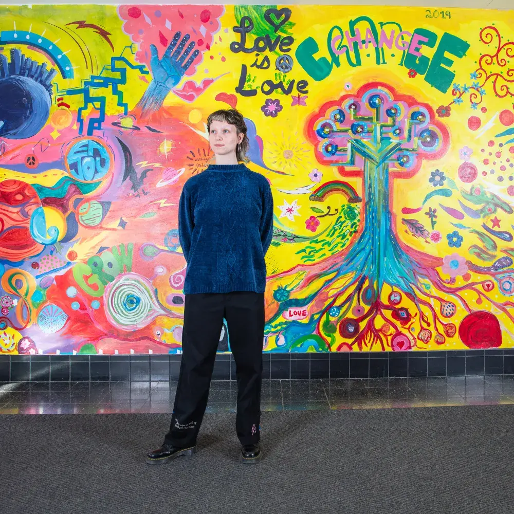 person in blue sweater and black pants in front of a colorful wall