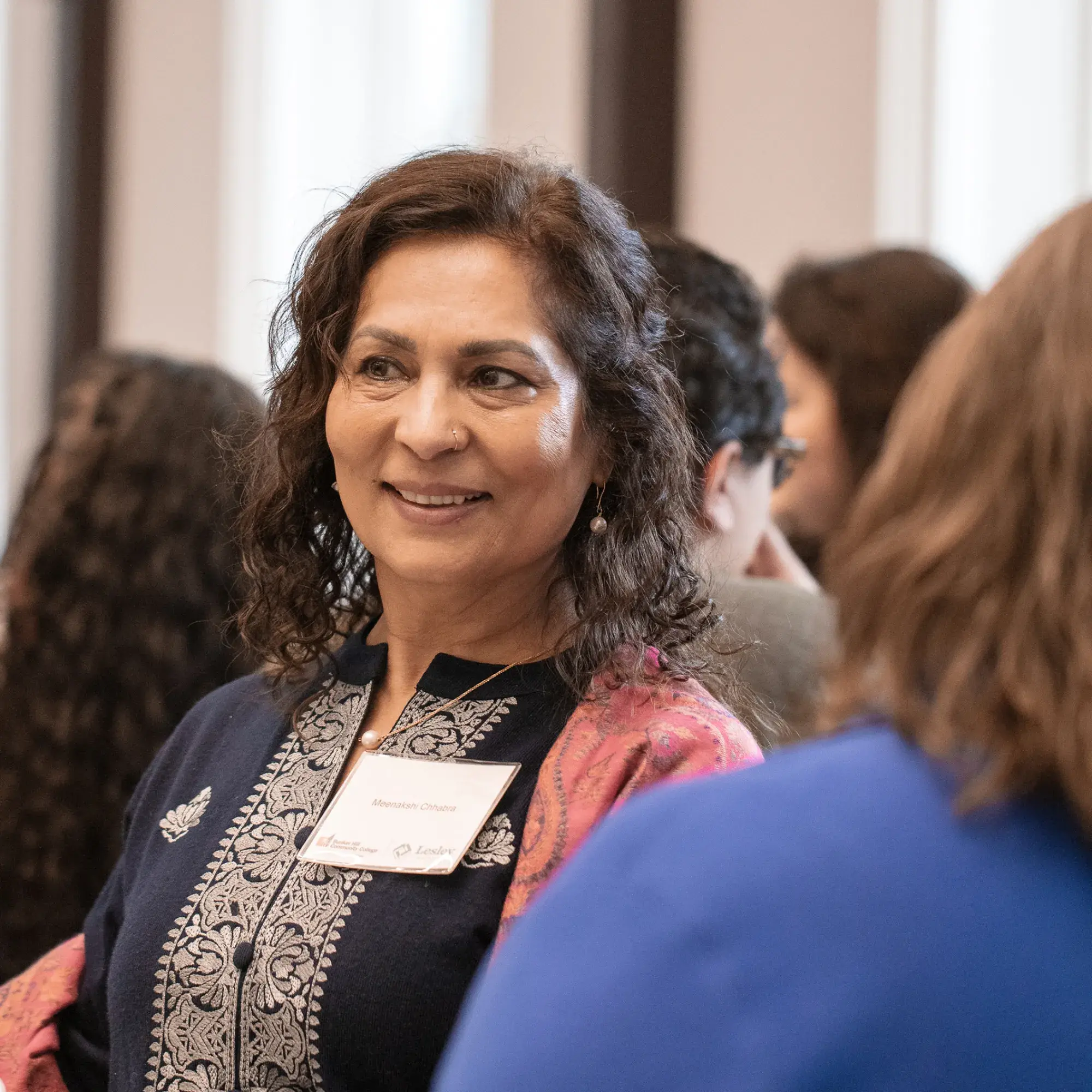 A smiling woman with dark, wavy hair stands among a group of people, looking to her left. She is wearing a dark blue garment with intricate white embroidery and a pink patterned shawl over one shoulder. A nametag that reads "Monisha Perona" is pinned to her chest. A green glass bottle is visible on a white surface in the foreground on the left. Other blurred figures are in the background.