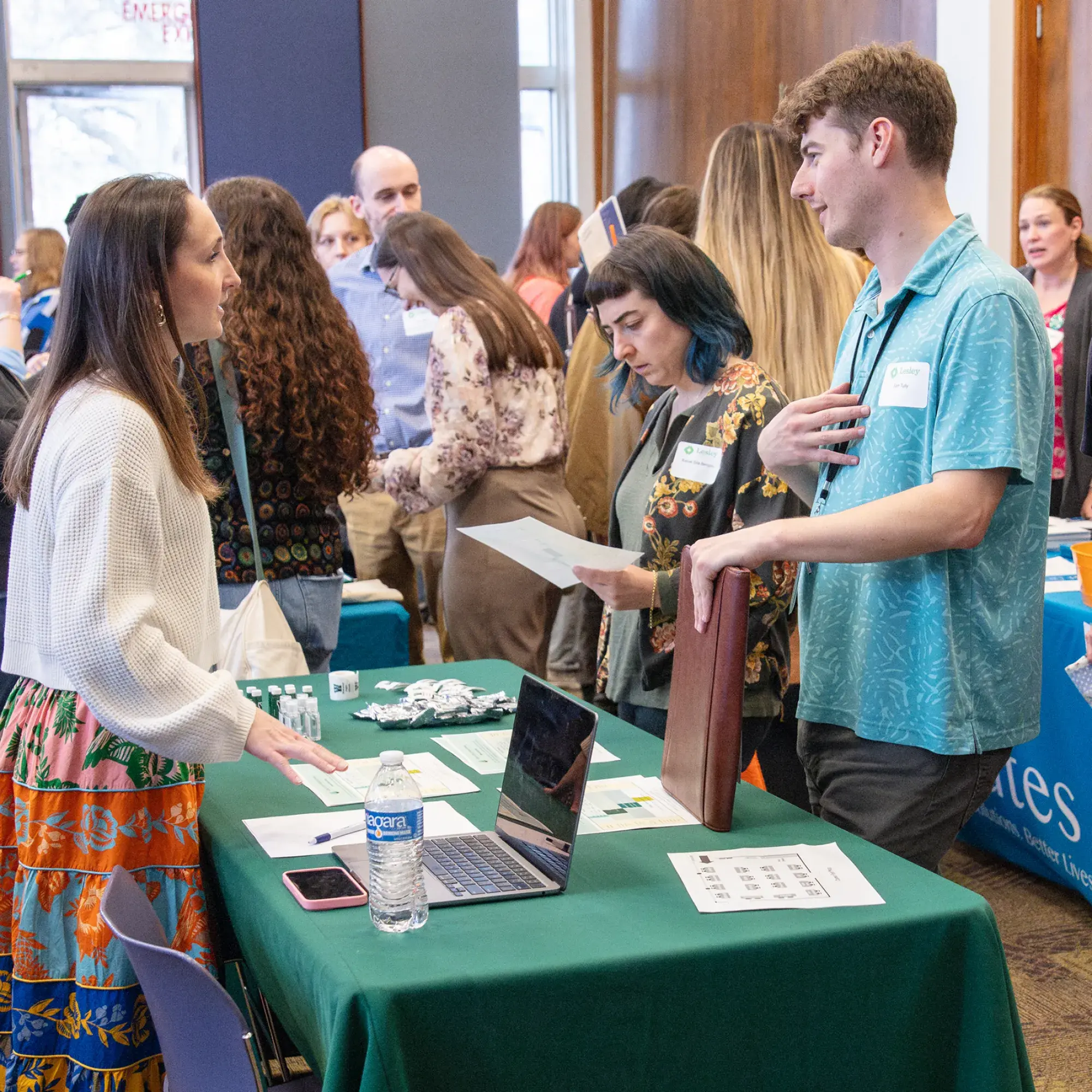 A large indoor event, likely a job fair, with numerous people milling about. In the foreground, a woman with dark hair, wearing a white sweater and a long, colorful floral skirt, stands on the left side of a green-draped table and is talking to a young man on the right. The man has short, light brown hair, wears a light blue patterned short-sleeved shirt, and holds a dark folder. A laptop, water bottle, and informational materials are on the table. In the background, many other attendees are visible.