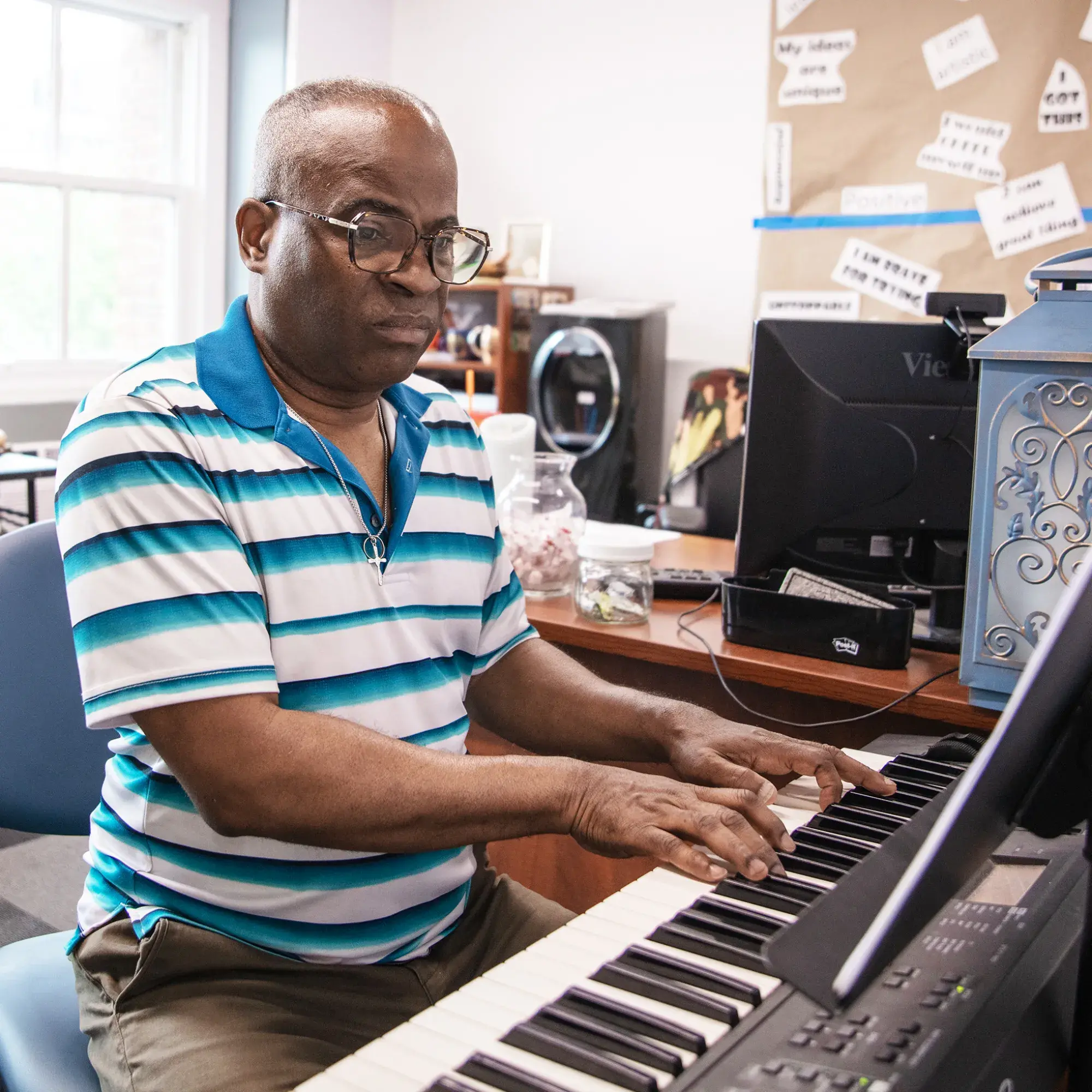 A man wearing glasses and a blue-striped polo shirt sits at an electric keyboard, playing music in a colorful and inviting room. The background features various musical instruments, including drums, and a bulletin board with motivational phrases. The space feels creative and supportive.